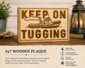 Wooden plaque with tugboat design and 'Keep On Tugging' text on a wooden surface.